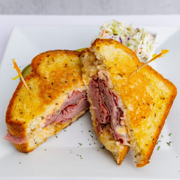 A Goldies Restaurant Reuben sandwich, cut in half, with coleslaw and a pickle spear on a white plate.