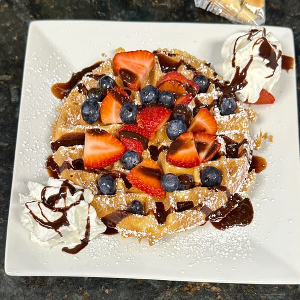 Golden waffle topped with strawberries, blueberries, chocolate drizzle, powdered sugar, and whipped cream.