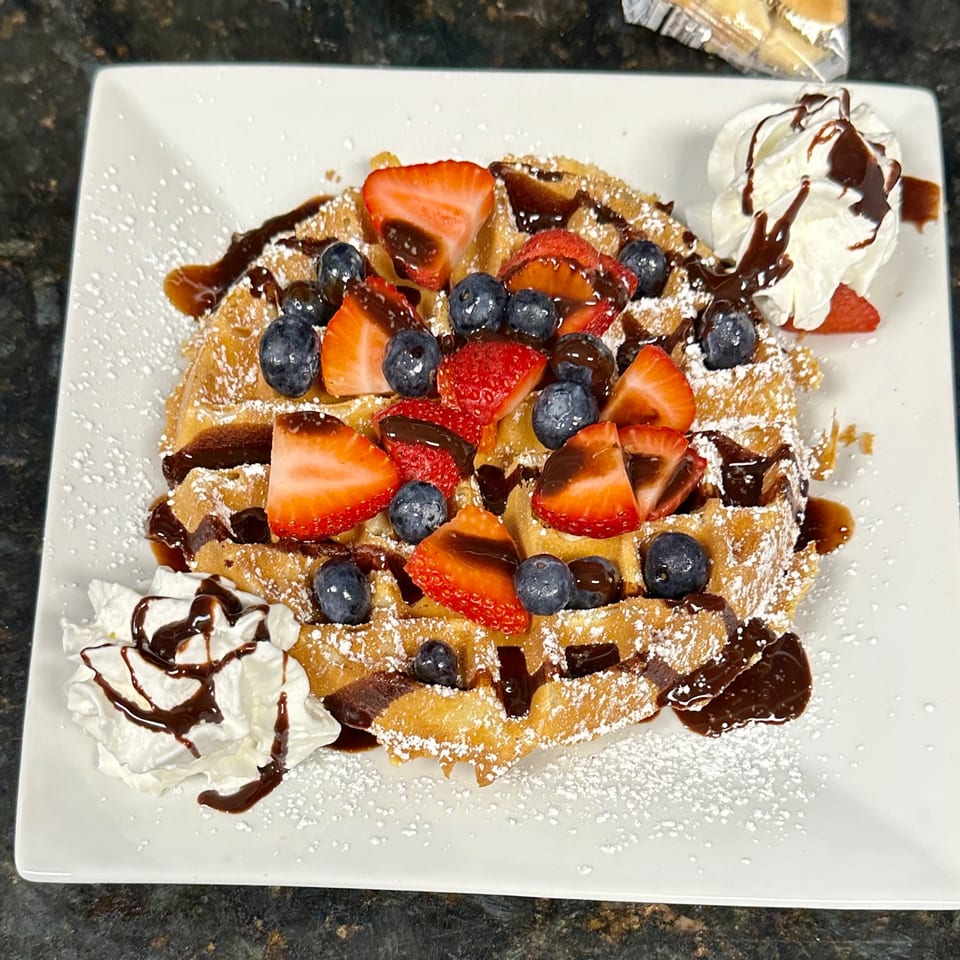 Golden waffle topped with strawberries, blueberries, chocolate drizzle, powdered sugar, and whipped cream.